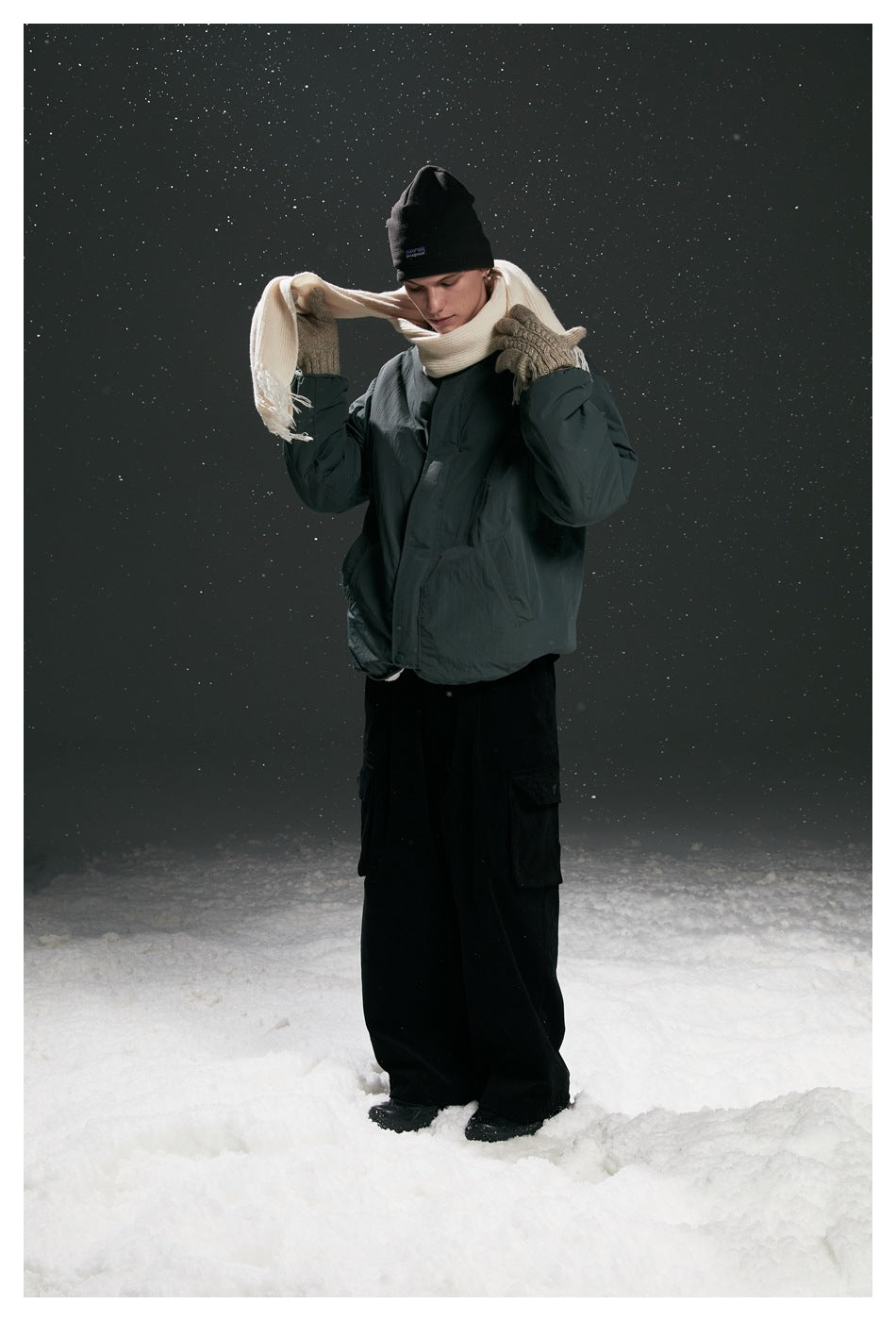 Model in dark green jacket, black beanie, cream scarf, gloves and black cargo pants standing in snow