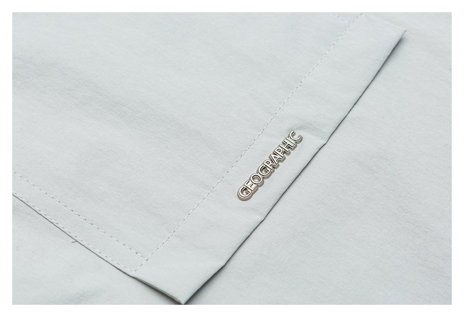 Close-up of a metal GEOGRAPHIC badge beside stitched seams on pale gray-green shirt fabric.