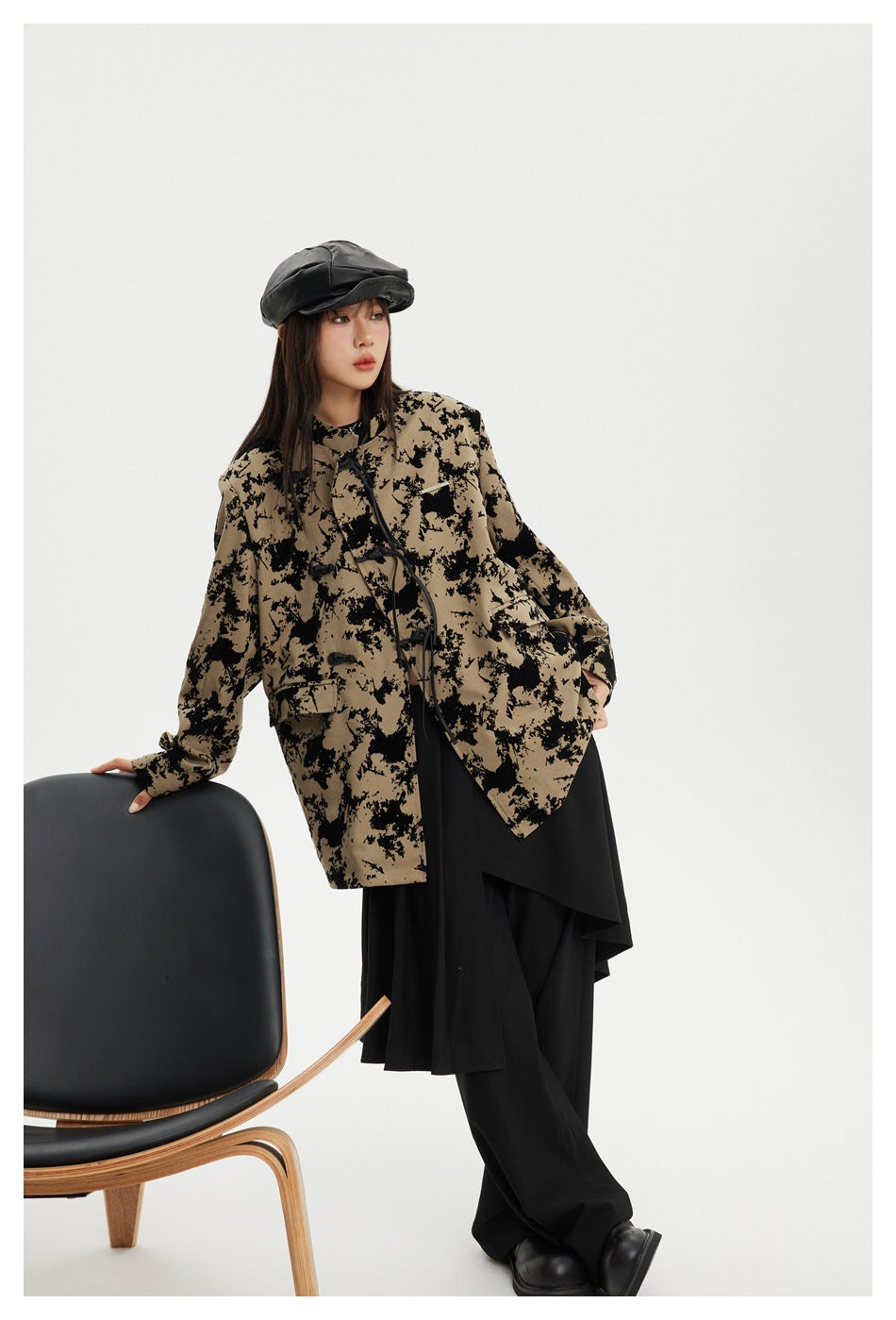 Full-length model wearing a tan-and-black patterned jacket with black layered trousers and a black cap.