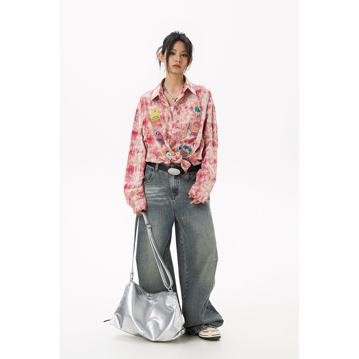Model wearing a pink marbled button-up shirt with colorful cartoon patches, tied at the waist, with wide-leg jeans.