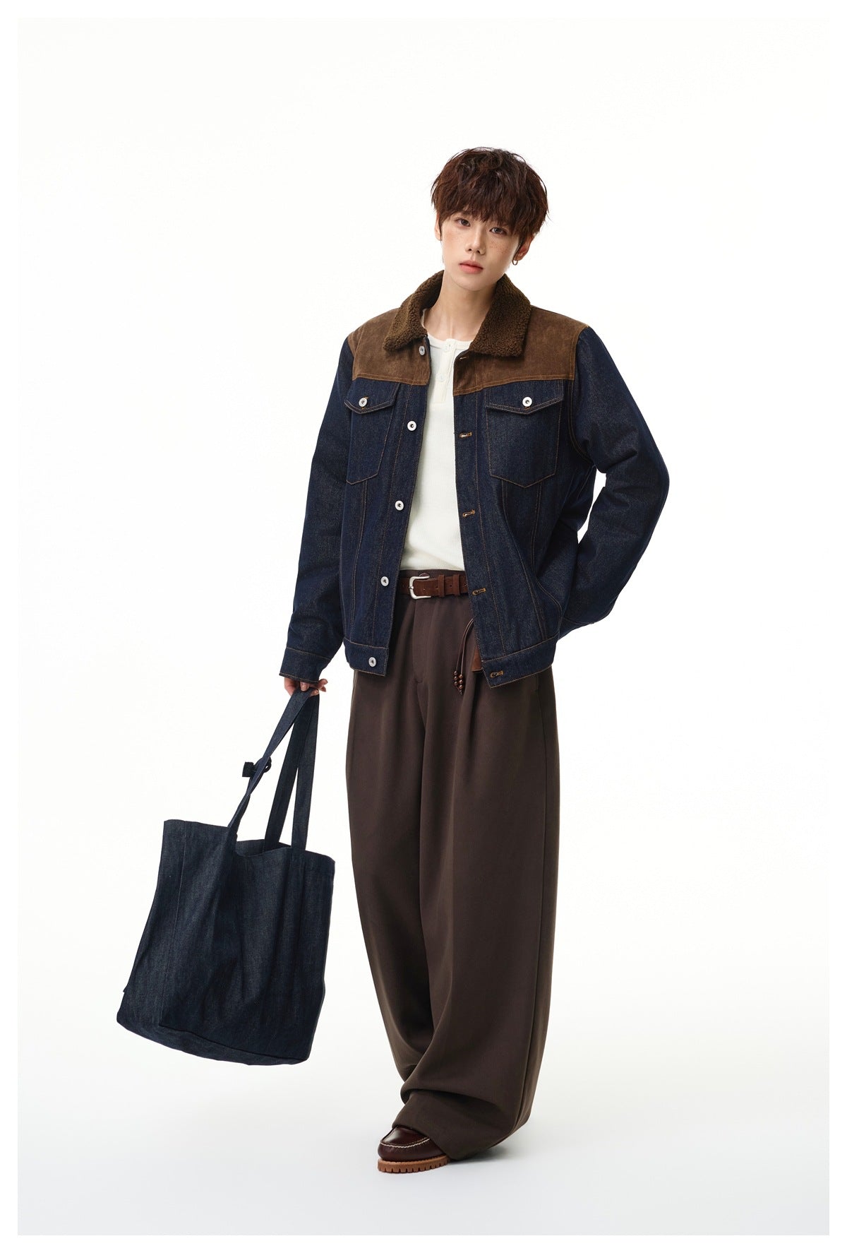 Model standing in a denim jacket with brown yoke and fleece-like collar, brown wide pants and holding a denim tote bag