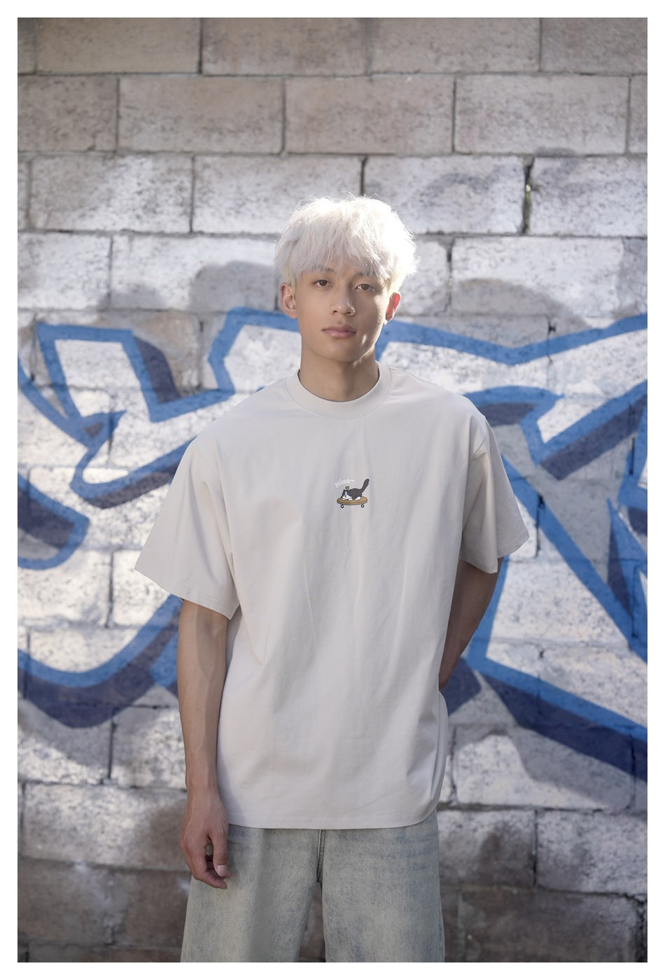 Model wearing a light beige oversized T-shirt with a small cat-on-skateboard chest graphic, against a stone wall.