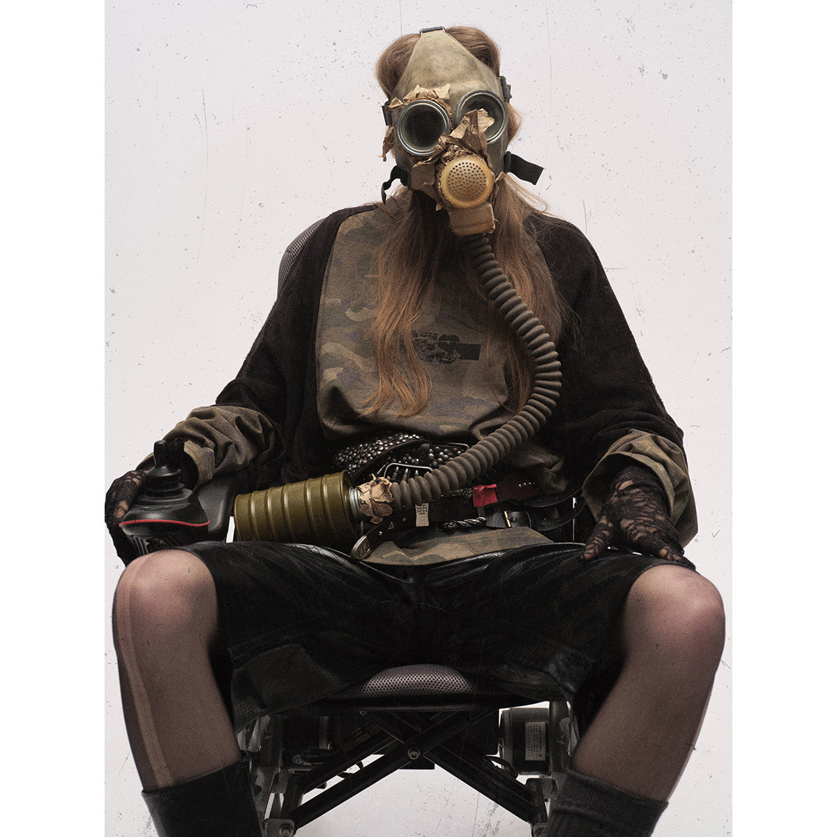 Seated model wearing an olive camouflage raglan top with black sleeves and black shorts, styled with a gas mask.