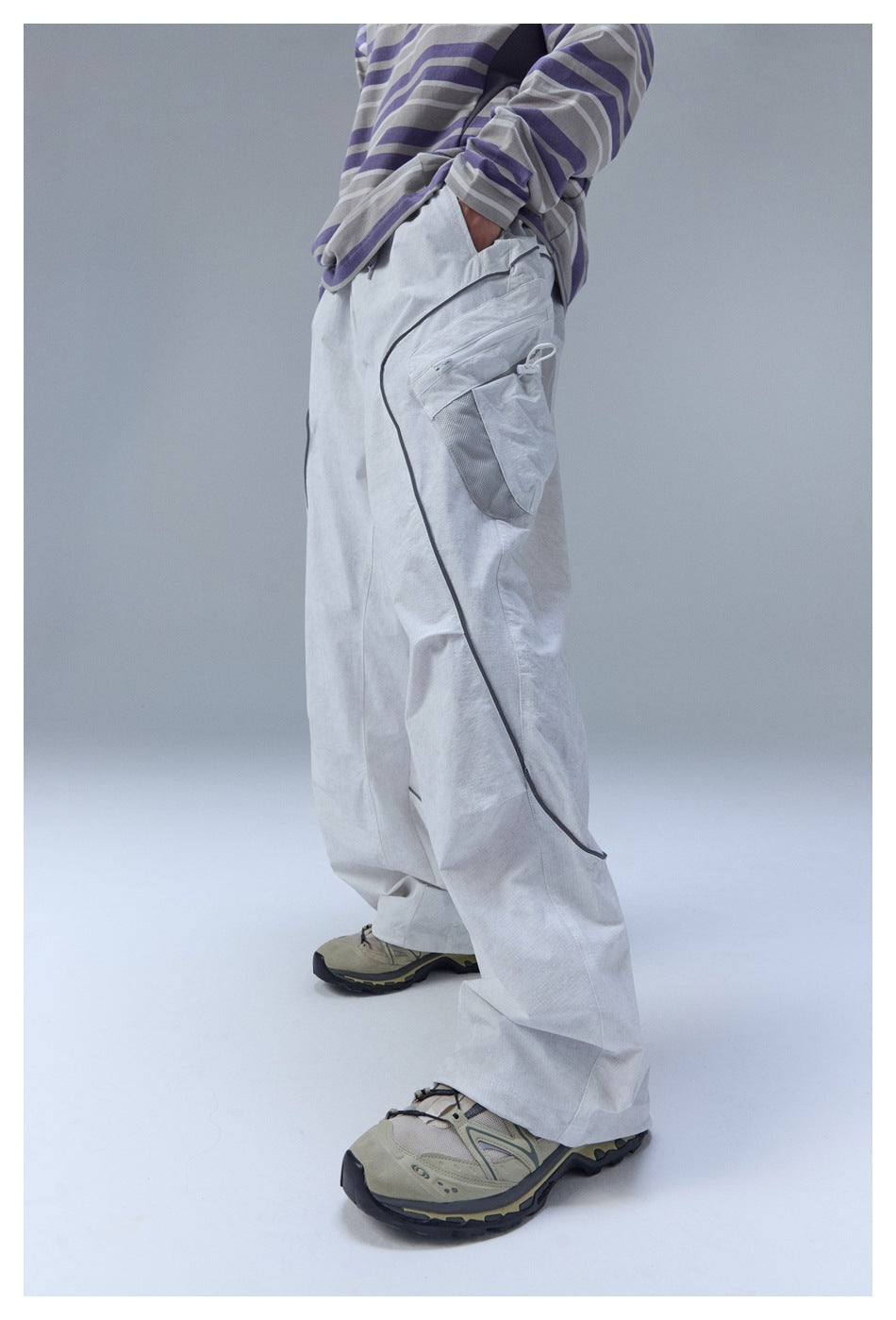 Side view of a model wearing loose light gray cargo pants with dark piping and utility pockets.