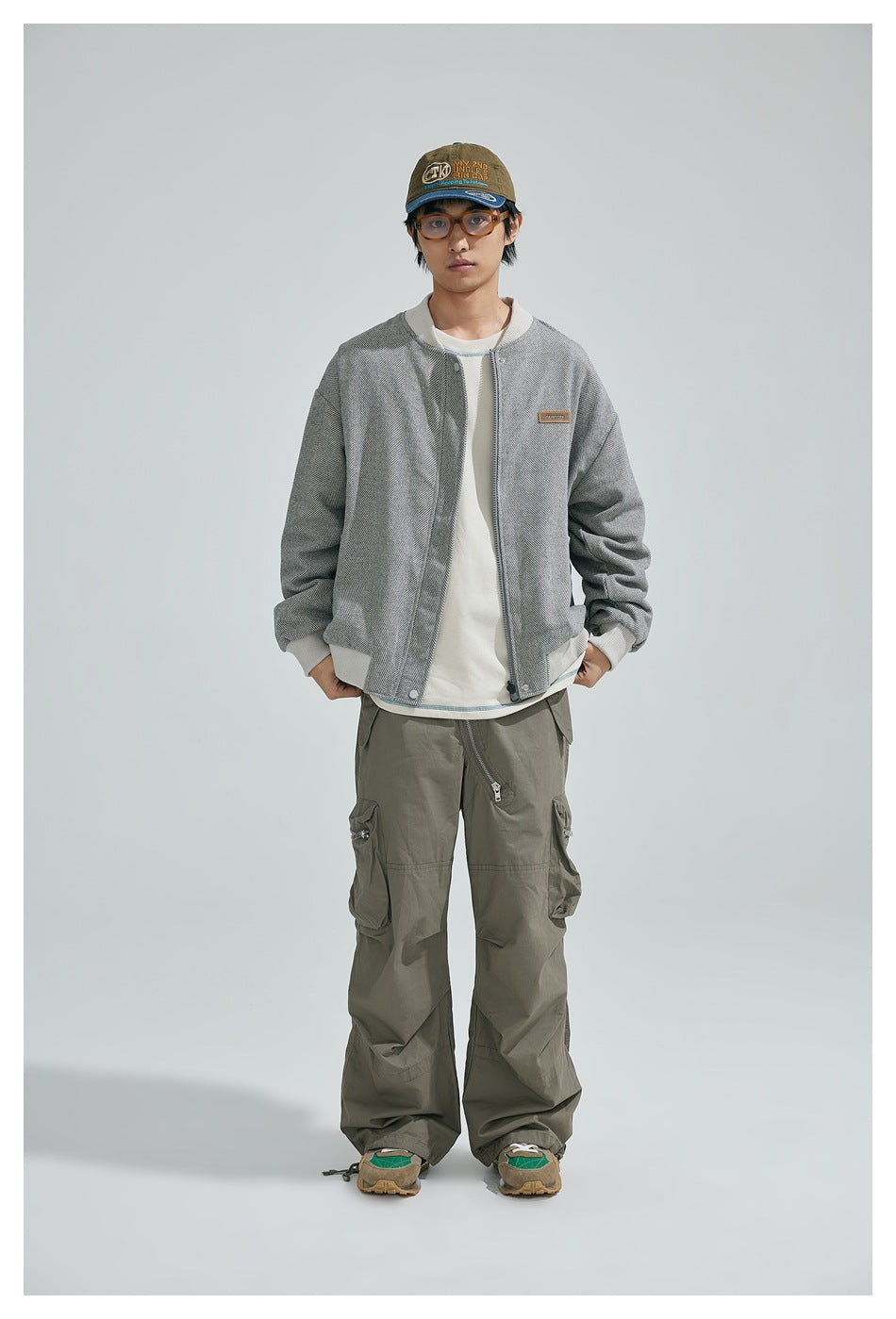 Model wearing gray herringbone bomber jacket with beige ribbed trim over a cream tee and olive cargo pants
