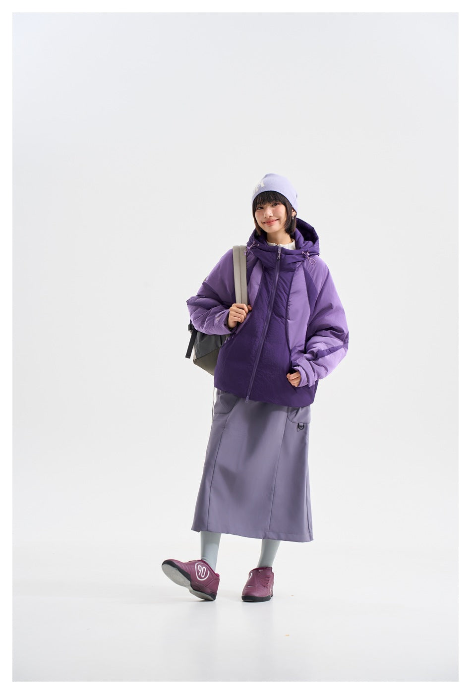 Model wearing a color-blocked purple hooded jacket with a lavender beanie and long gray skirt.