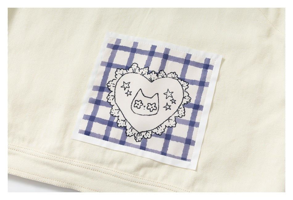 Close-up of an ivory garment with a blue-and-white checked patch featuring a lace-edged heart, stars, and a cat face.