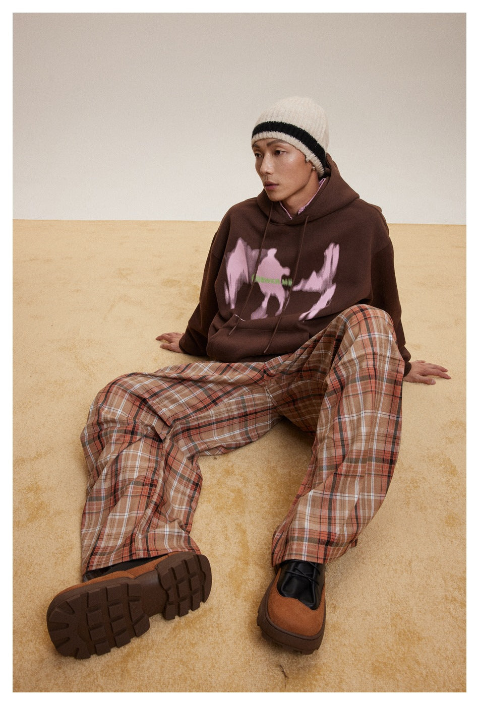 Seated model wearing a dark brown hoodie with a pink graphic, plaid trousers and a cream striped beanie.