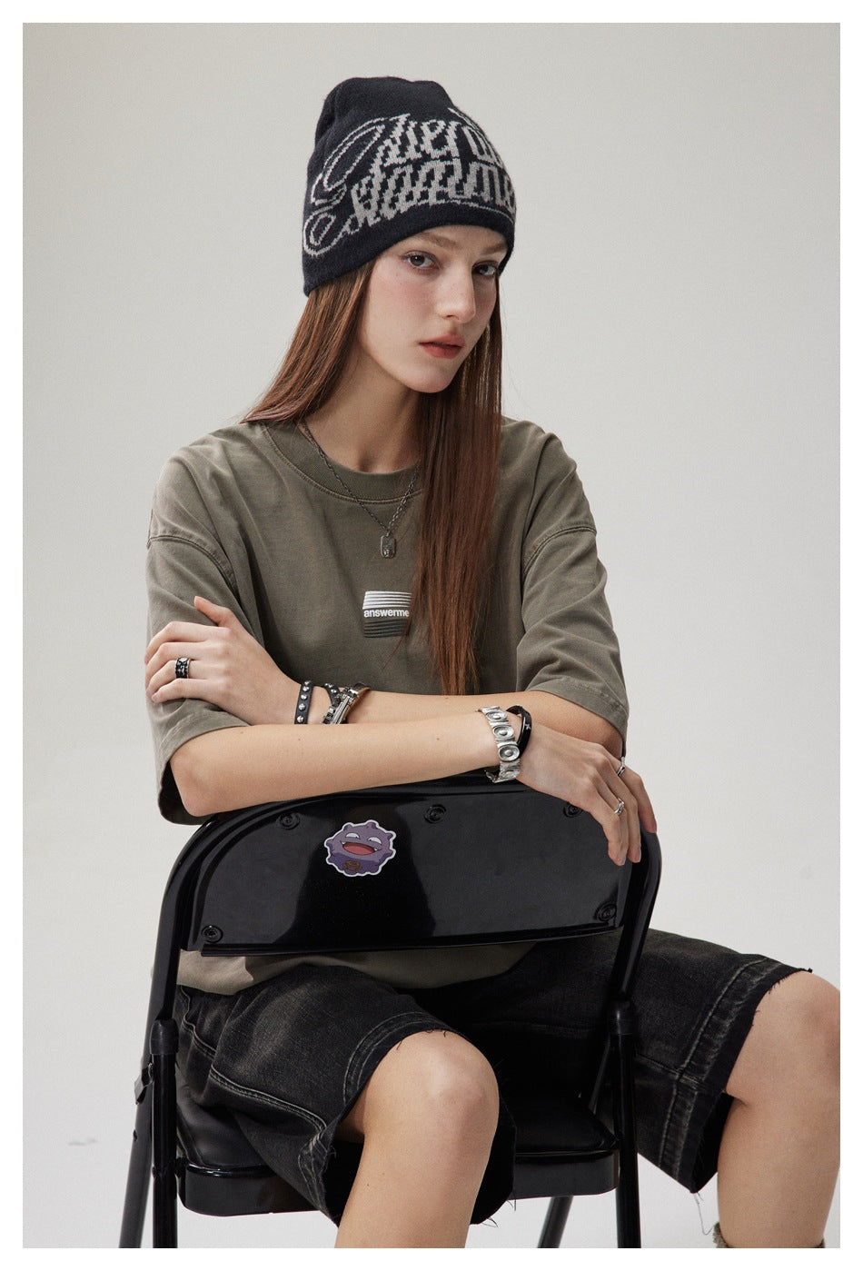 Model seated in an olive-gray oversized T-shirt with a small chest graphic and black denim shorts.