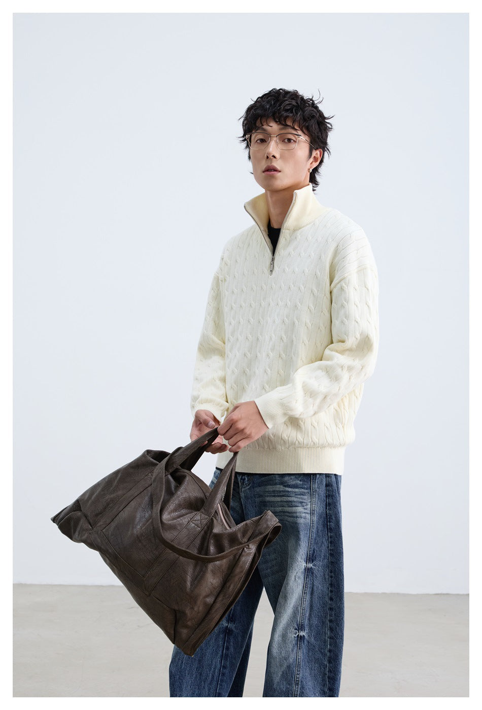 Model wearing an ivory cable-knit quarter-zip sweater and blue jeans, holding a dark brown tote bag.