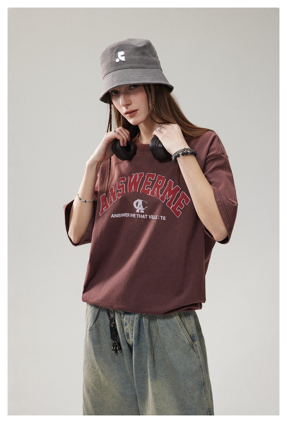 Model wearing a burgundy graphic T-shirt and gray bucket hat, holding black headphones at her shoulders.