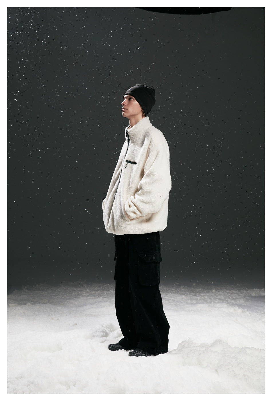 Side view of a model wearing a cream fleece zip jacket with dark trim and black cargo pants in falling snow.
