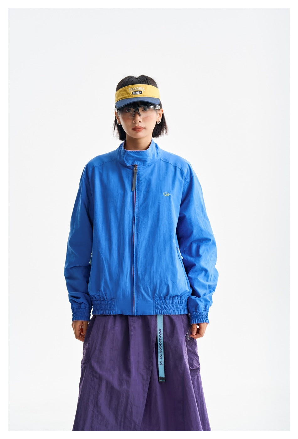 Model wearing a bright blue zip-front jacket with a high collar and elastic cuffs, paired with a purple skirt.