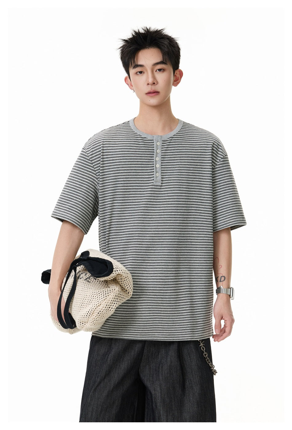 Model wearing gray striped short-sleeve Henley T-shirt with dark wide pants, holding a cream mesh bag