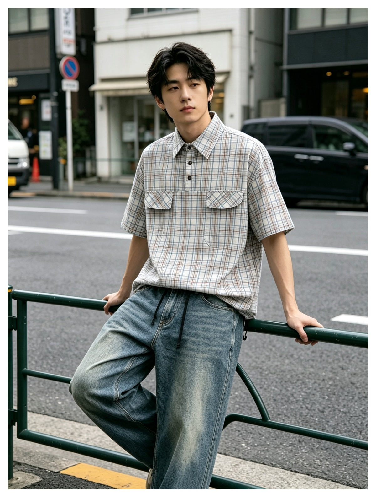 Man in a beige plaid short-sleeve shirt with chest flap pockets and wide-leg jeans leaning on a street railing
