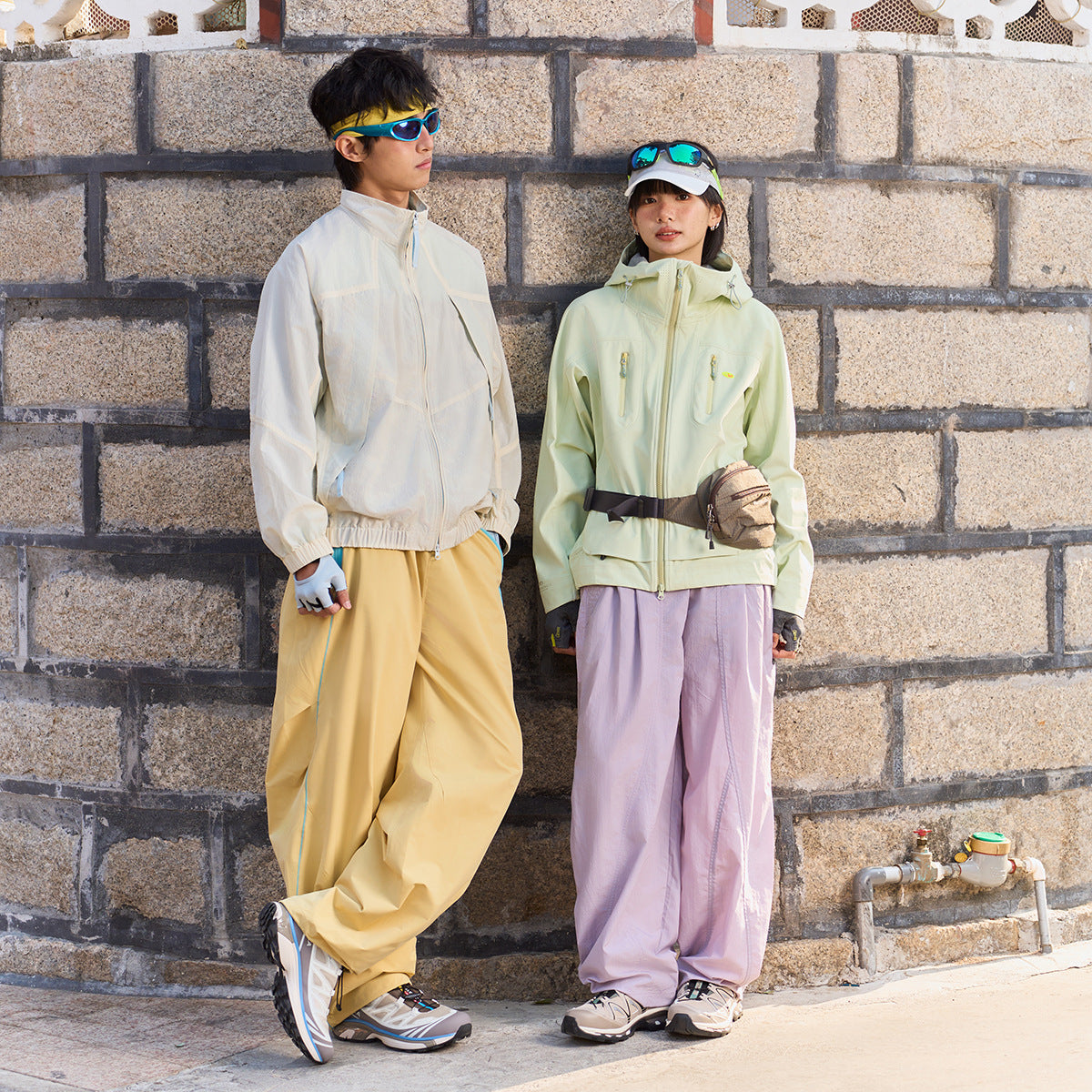 Two models wear a cream zip jacket and pale green hooded jacket with loose yellow and lavender trousers.