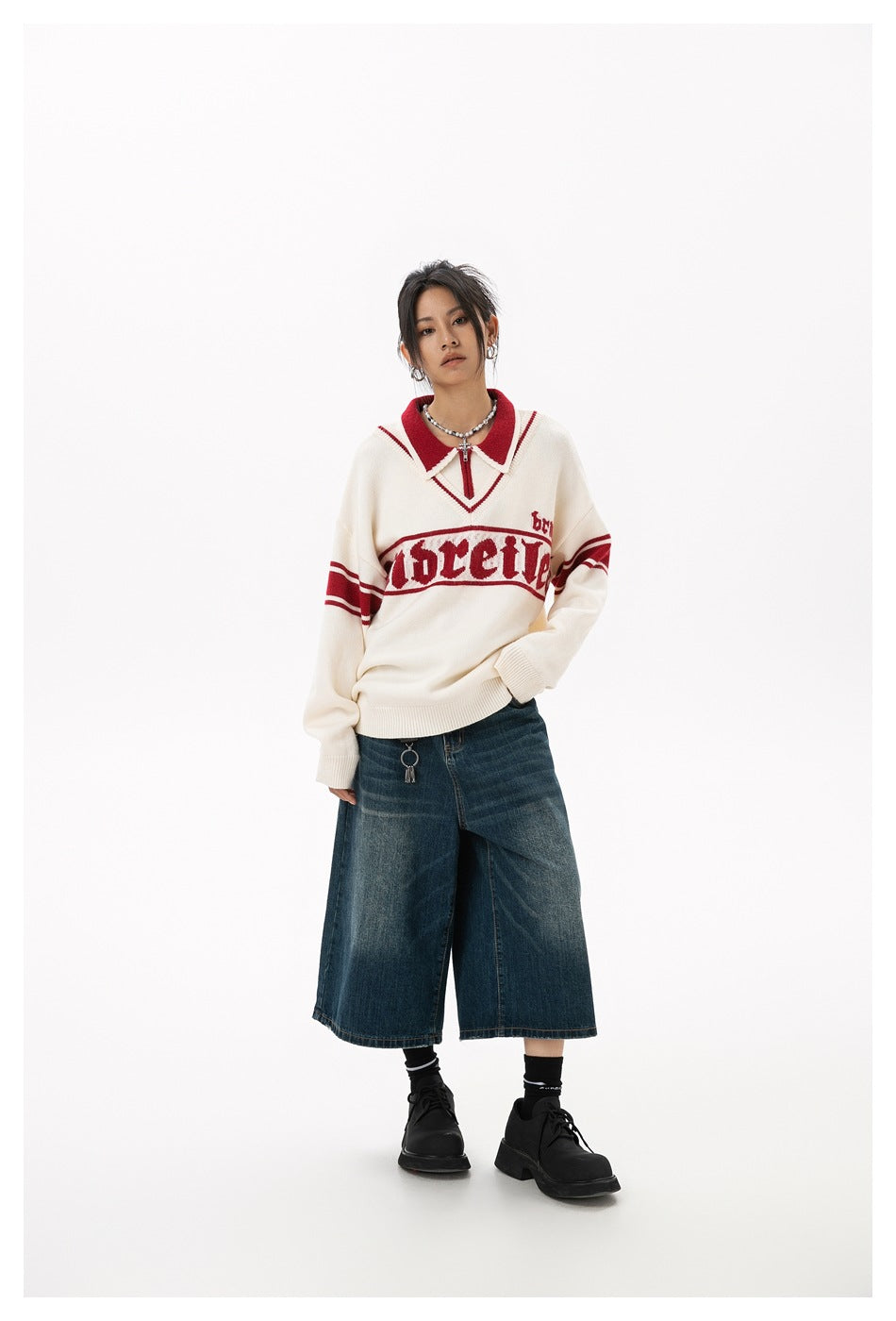 Model wearing an ivory sweater with red trim and lettering, styled with wide-leg denim shorts and black shoes.