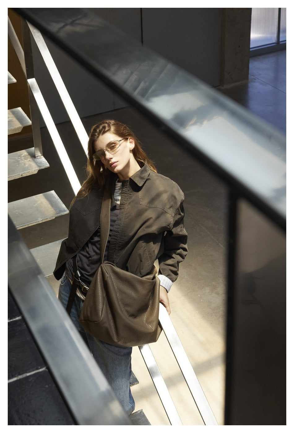 Woman on a staircase wearing an olive-brown zip jacket over a dark top with jeans and a large shoulder bag