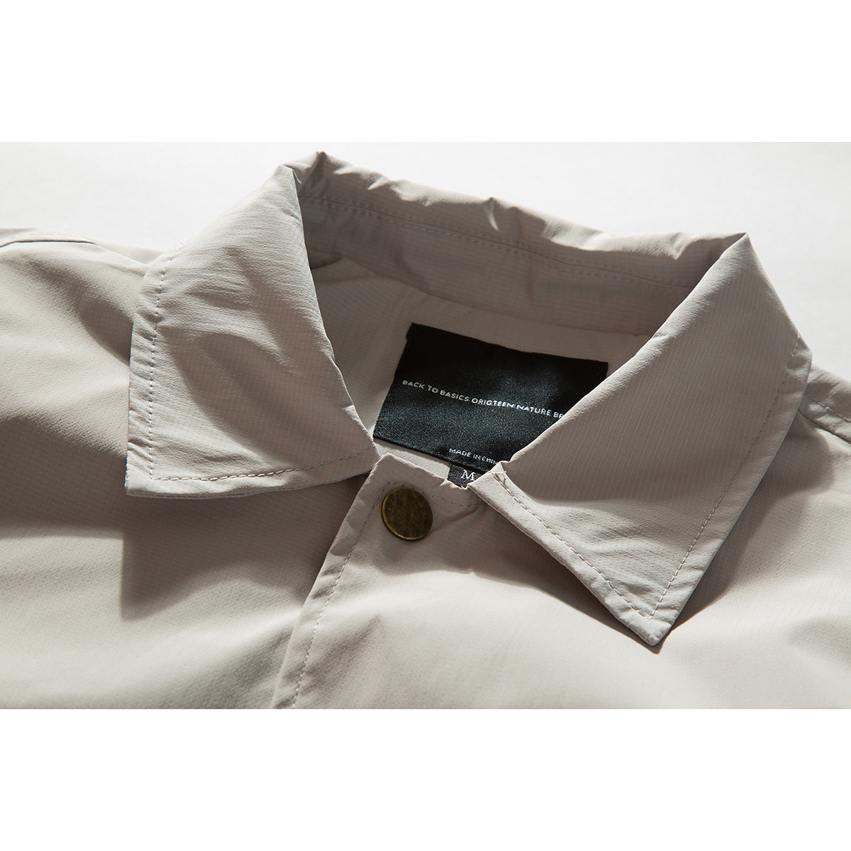 Close-up of a light beige jacket collar, showing a brass-tone button and black inner neck label.