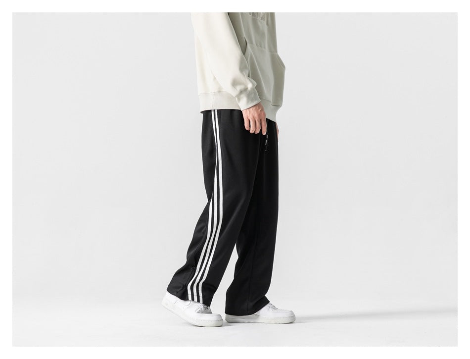 Side view of a cropped model wearing loose black track pants with three white side stripes.