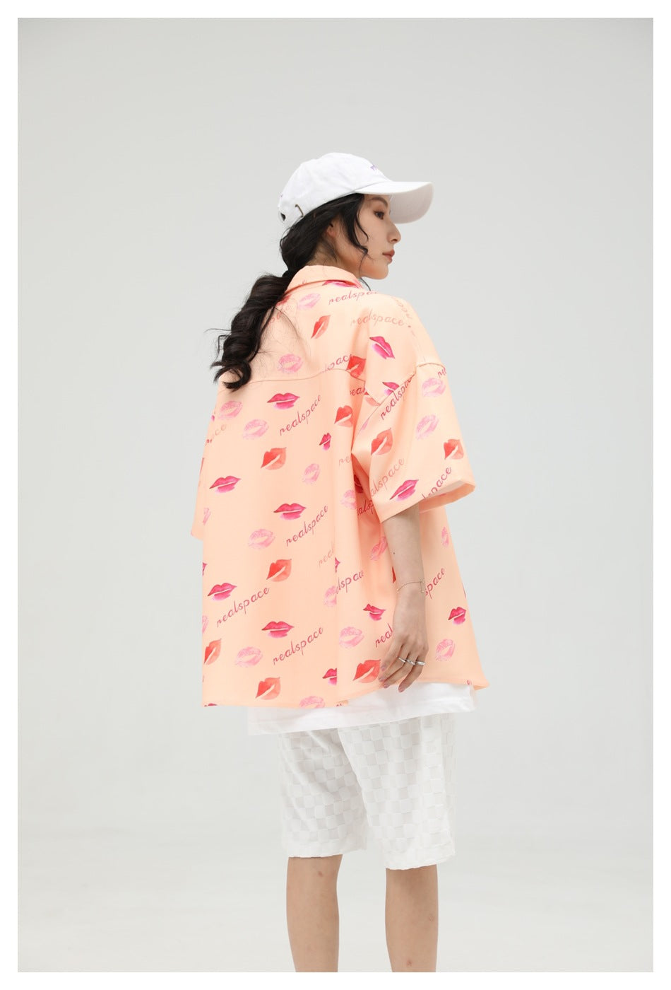 Back view of a model wearing the peach lip-print oversized short-sleeve shirt with white shorts and a white cap