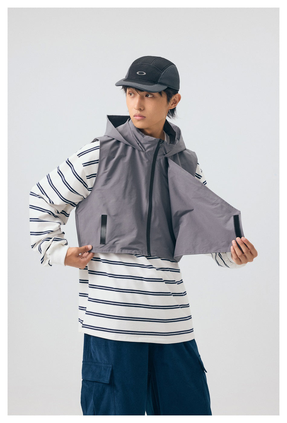 Model displays a gray hooded vest open over a white striped shirt, with navy cargo pants and a black cap.
