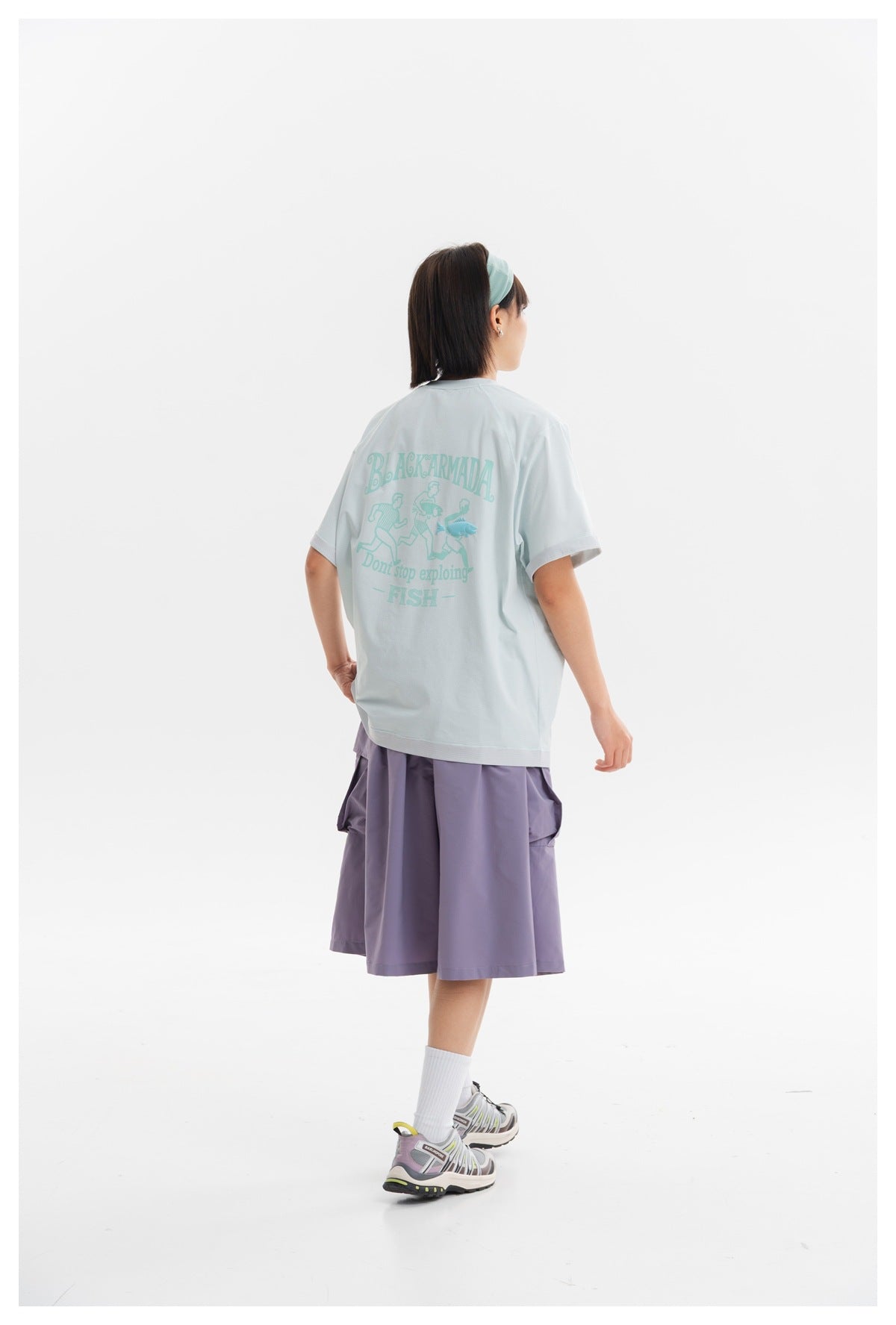 Rear view of a model wearing a pale aqua graphic T-shirt and loose lavender cargo shorts.