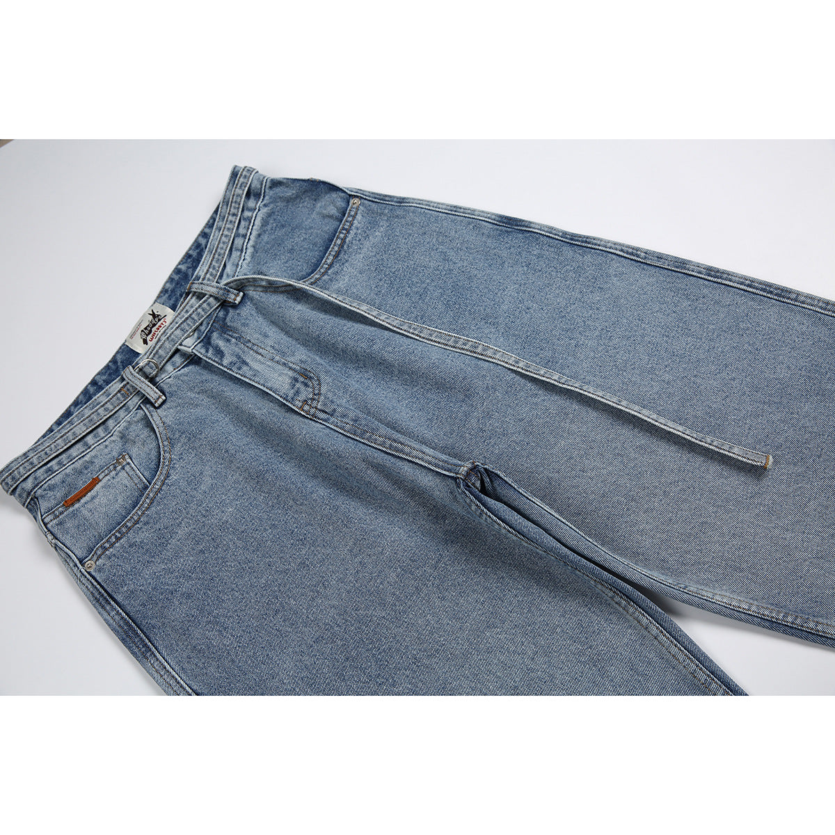 Close-up of light blue jeans laid diagonally, showing the front pocket, waistband, belt loops and long hanging belt.