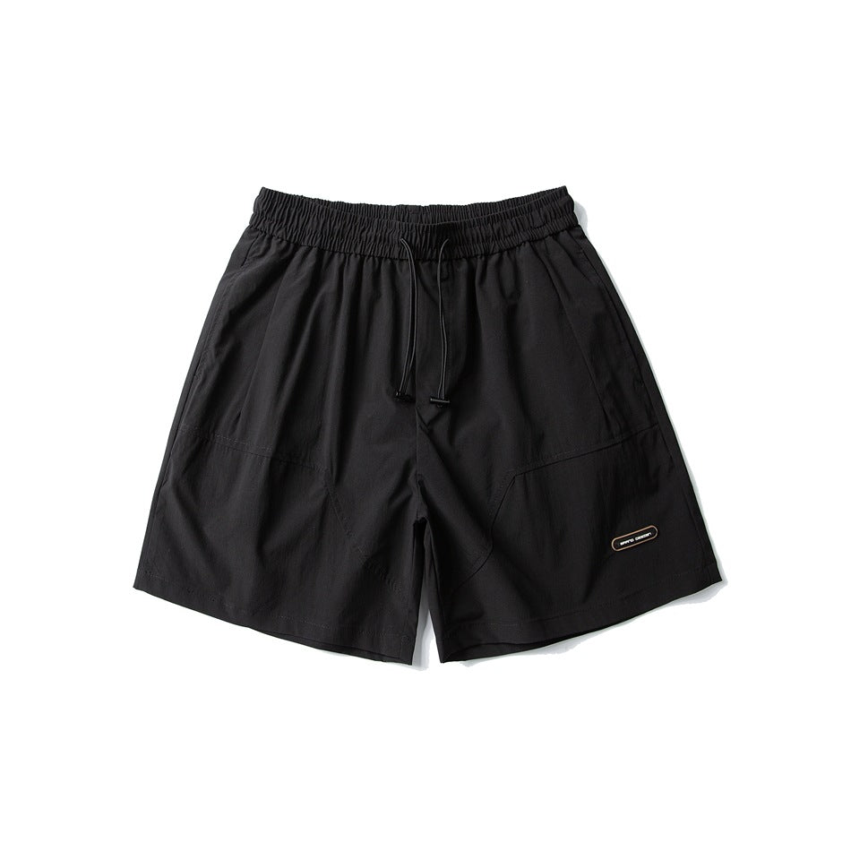 Black shorts laid flat, front view, with an elastic drawstring waist, angled seams, and a small leg patch.