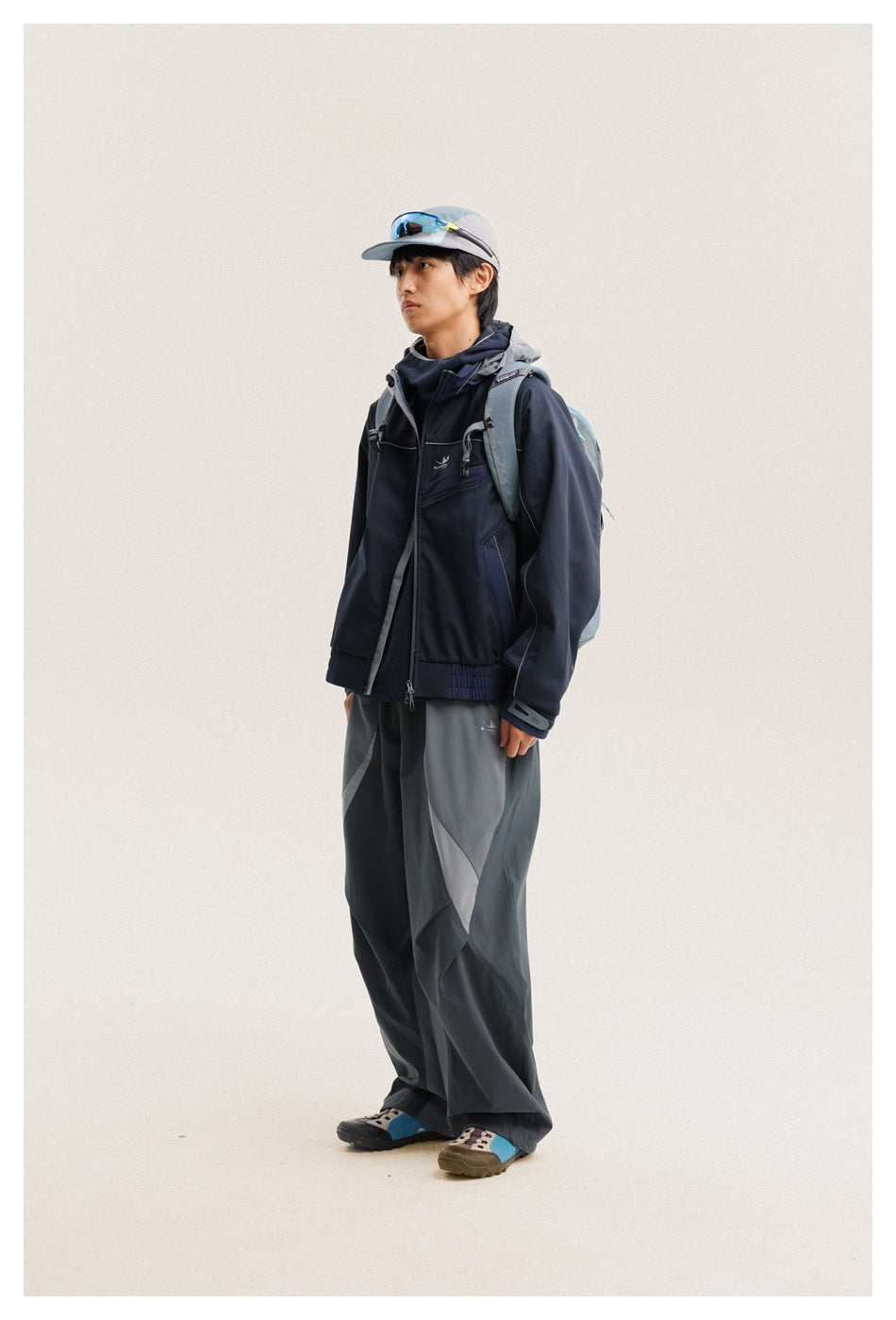 Full-length view of a model in a navy hooded jacket and loose gray paneled trousers, wearing a cap and backpack.