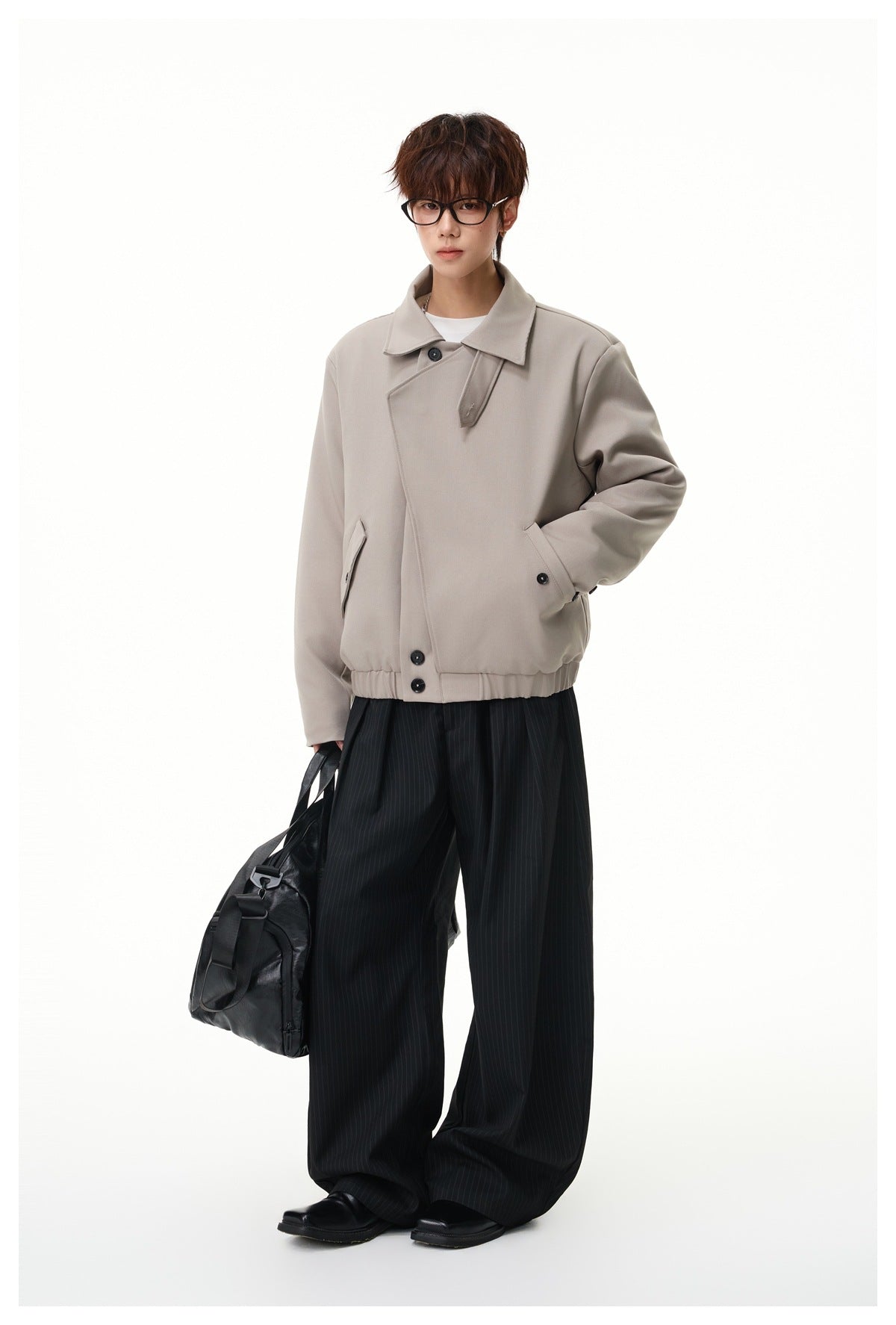 Model wearing a gray khaki short jacket with wide pinstripe trousers and holding a black duffel bag