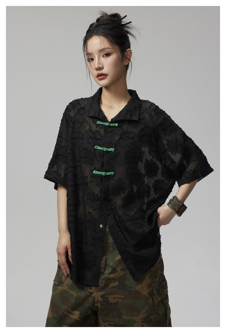 Front view of a model wearing a black sheer floral-textured shirt with green frog closures and camouflage trousers.