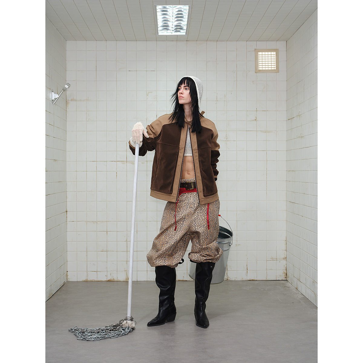 Model wearing a two-tone brown zip jacket and leopard-print trousers, holding a mop in a tiled room.