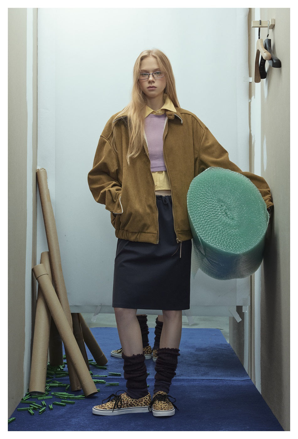 Model wearing a brown corduroy-style zip jacket over a lilac top and dark skirt, holding a roll of bubble wrap