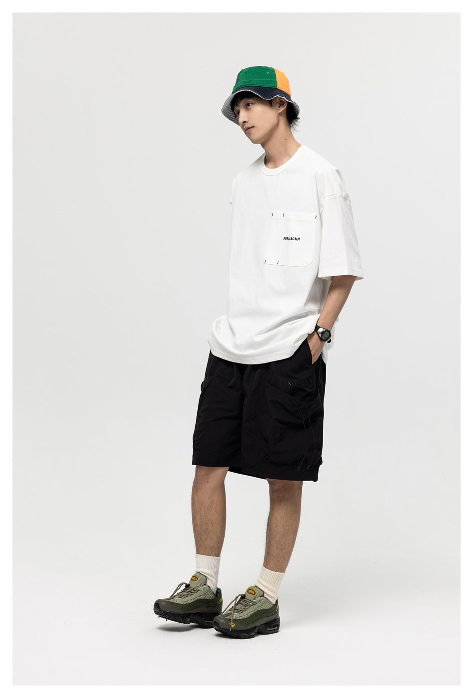 Model in a white oversized pocket T-shirt with black shorts and a colorblock bucket hat, angled pose with hand in pocket
