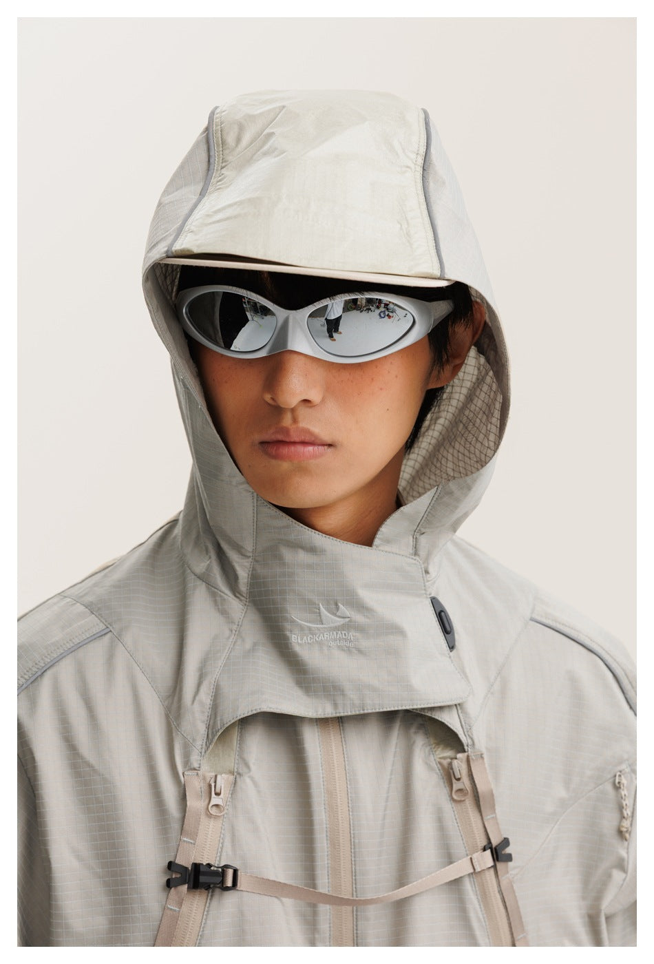 Close-up of a model wearing a light gray hooded shell jacket with a high collar, zip pockets and chest straps.