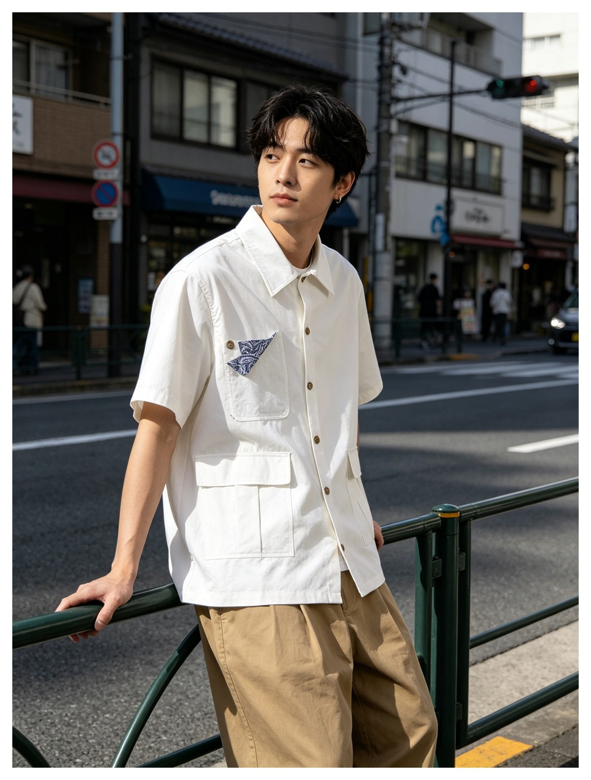 Man wearing white short-sleeve pocket shirt with khaki trousers, leaning on a green railing on a city street