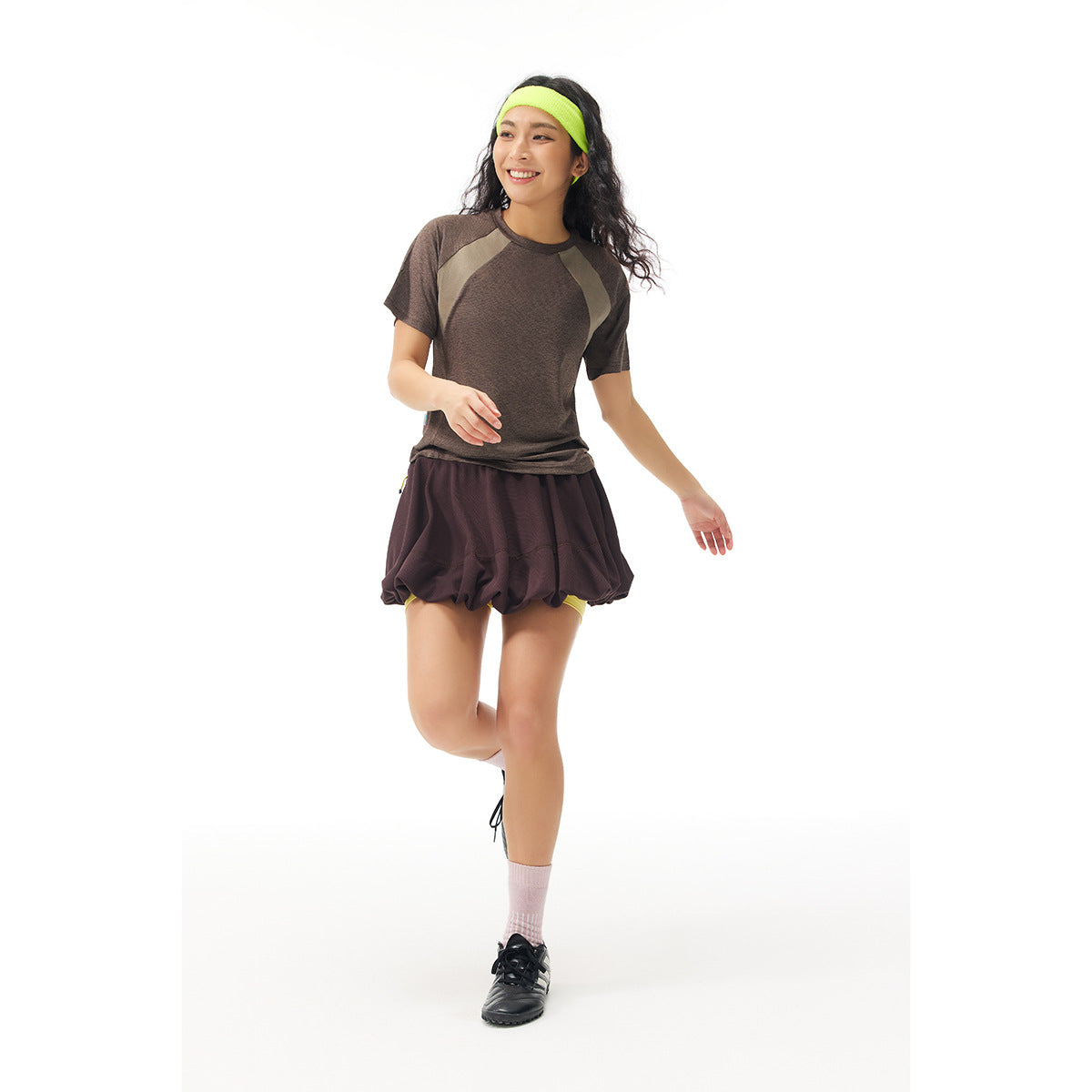 Model wearing a brown color-block athletic T-shirt and dark plum gathered shorts with a neon yellow headband.