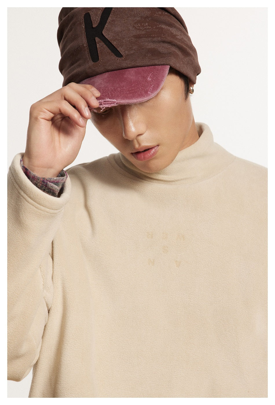 Close-up of a model in a cream high-neck pullover and brown cap with a distressed pink brim.