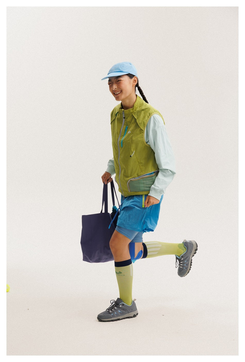 A model wears a lime-green sleeveless vest over a pale mint top, blue shorts, and a light blue cap, shown from the side.