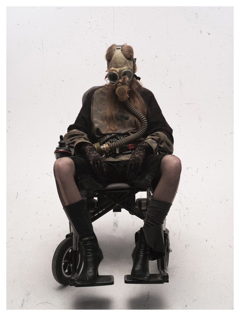 Seated model wearing an oversized brown camouflage raglan shirt with black sleeves, layered belts, shorts and tall boots.