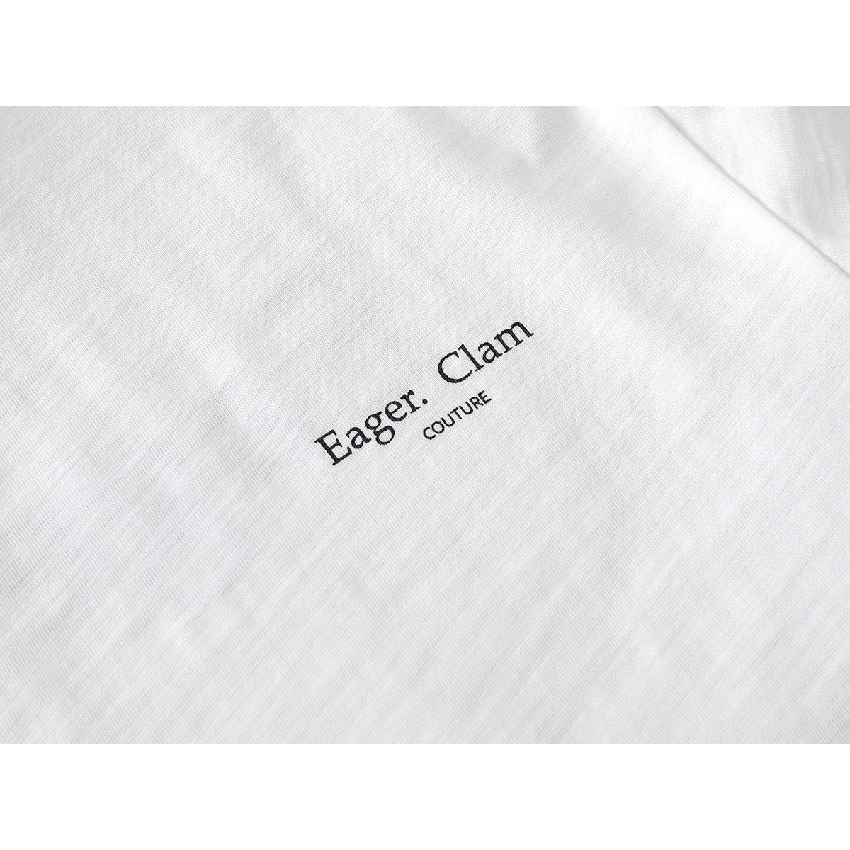 Close-up of a white shirt with black “Eager. Clam COUTURE” lettering on the chest.