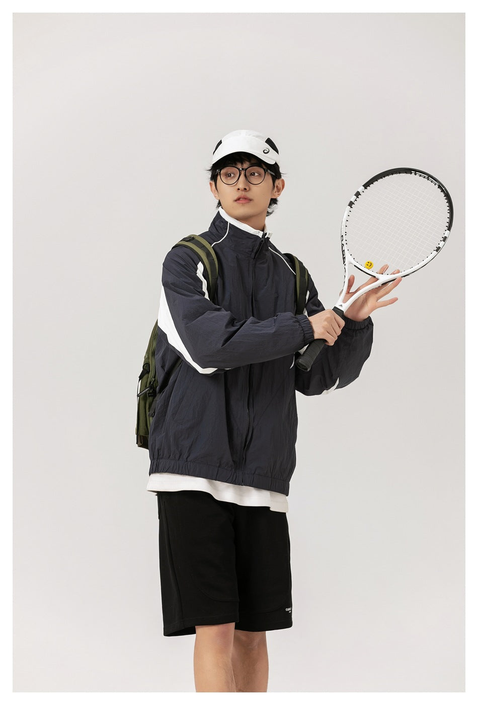 Model wearing a dark navy zip jacket with white panels and black shorts, holding a tennis racket.