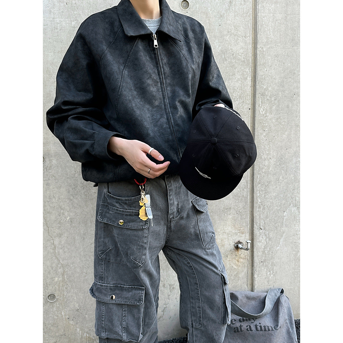 Model wearing a dark charcoal zip-front jacket with gray cargo pants and holding a black cap.
