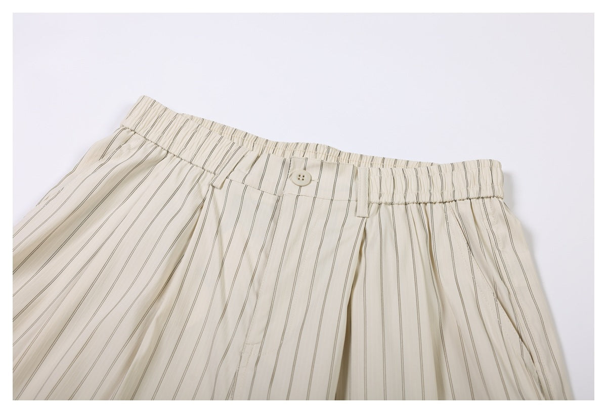 Close-up of the front waistband and pleats on cream shorts with thin dark vertical stripes and a button closure.