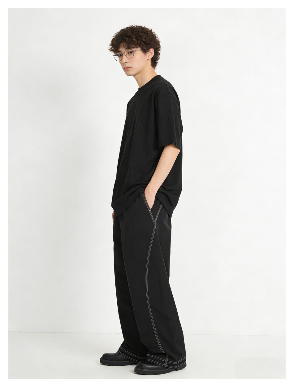 Model in a black T-shirt and black wide-leg pants with white contrast stitching, hand in pocket, side view