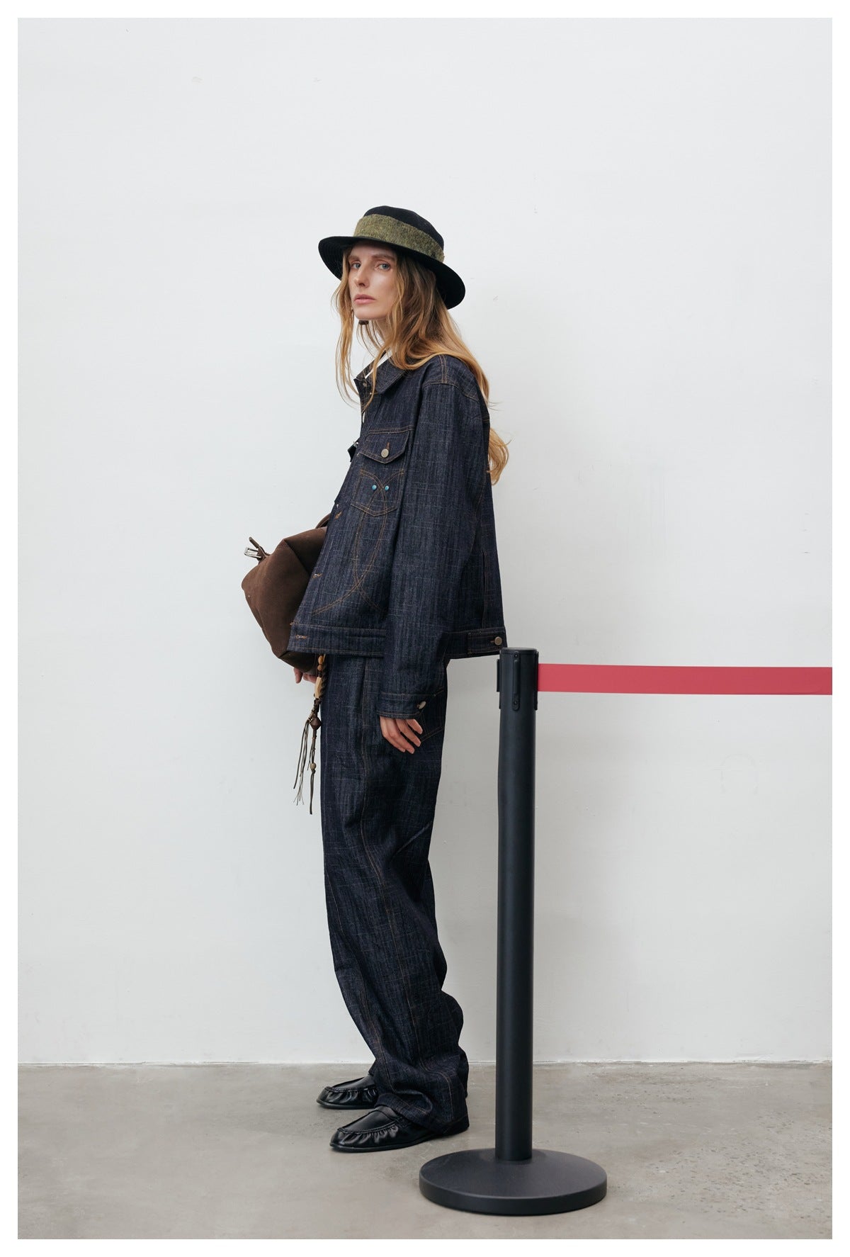 Model in side pose wearing a cropped dark denim jacket with matching wide-leg denim pants, a hat and a brown bag