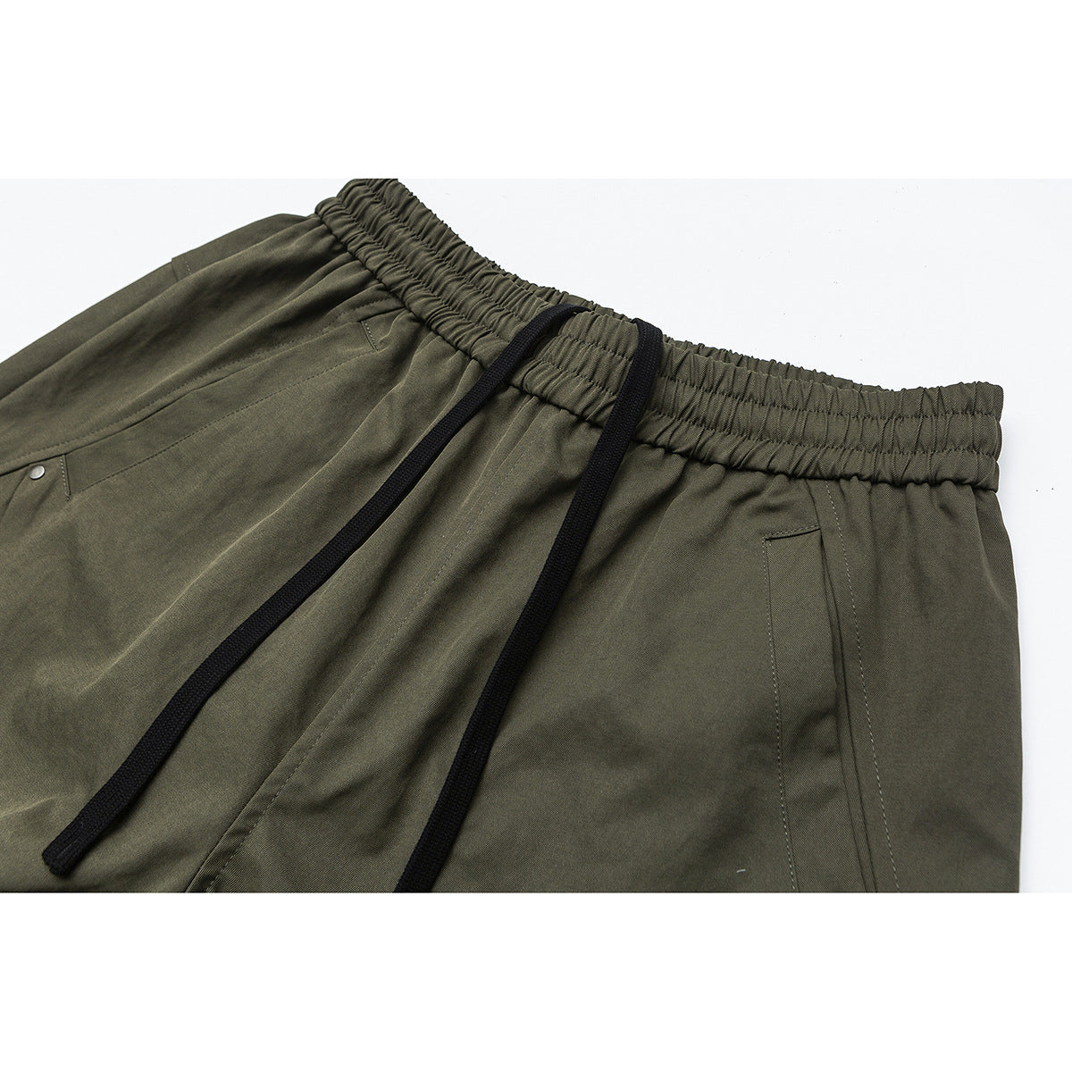 Close-up of olive trousers with a gathered elastic waistband, black drawstrings, and stitched pockets.