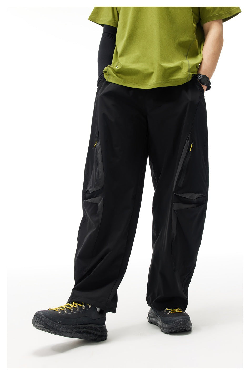 Model wearing loose black cargo pants with large thigh pockets and yellow zipper pulls, paired with hiking shoes.