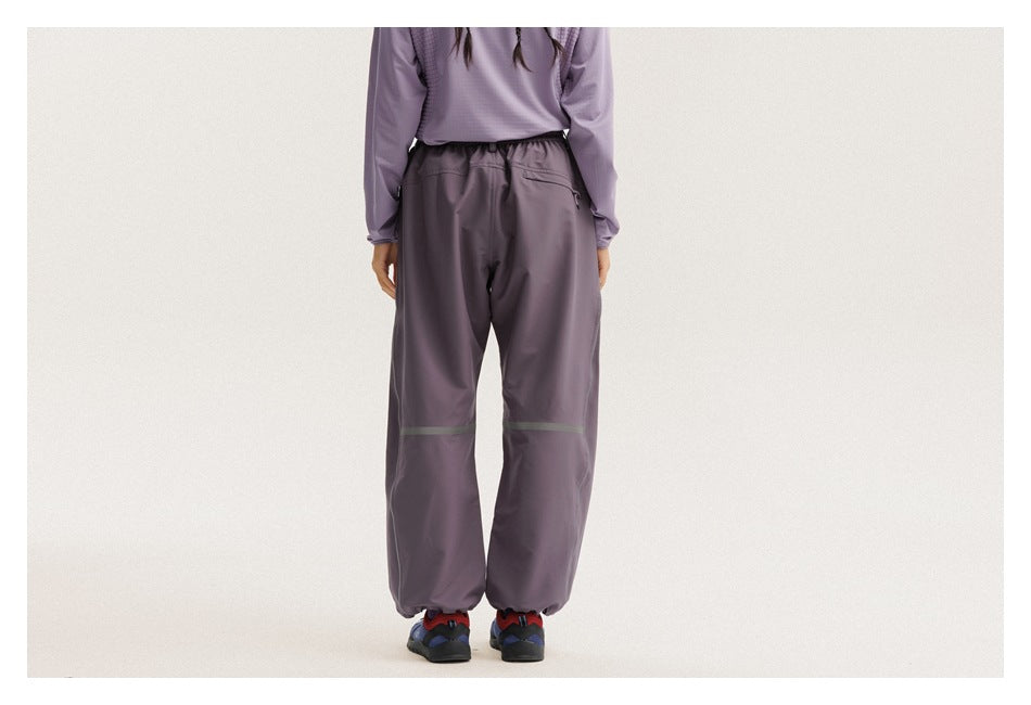 Rear view of a model wearing charcoal-purple loose pants with an elastic waist, back pocket, reflective bands and gathered
